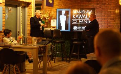 Zdjęcie ukazuje scenę, gdzie dwaj mężczyźni stoją na przeciwko siebie i rozmawiają. Widać również wyświetlacz oraz osoby siedzące przy stolikach, uczestniczące w wydarzeniu. 