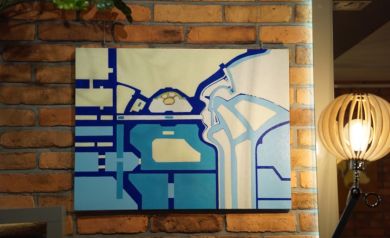 Na zdjęciu abstrakcyjny obraz wiszący na ceglanej ścianie. Obok stoi lampa. 