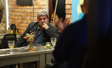 Mężczyzna ubrany w ciemną marynarkę siedzący przy stoliku. Podpiera sobie głowę ręką. Wpatruje się przed siebie. Wokół widać ceglane ściany. 