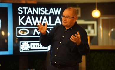 Na zdjęciu mężczyzna w średnim wieku, ma okulary. Gestykuluje rękoma i prawdopodobnie zwraca się do kogoś.  Za nim widać ciepłe światło.