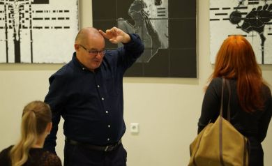 Mężczyzna i dwie kobiety stojący przy wiszących obrazach. Mężczyzna unosi jedna rękę do góry. 