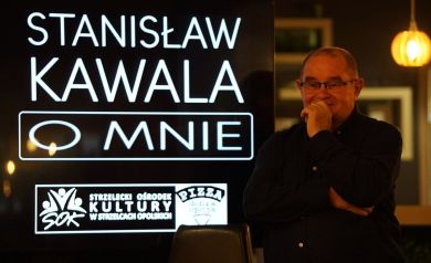 Mężczyzna ubrany w ciemną koszulę, w okularach podpiera ręką głowę. Obok niego wyświetlacz z napisem ,,Stanisław Kawala O MNIE', logiem Strzeleckiego Ośrodka Kultury i logiem pizzerii Dobra Opcja. 