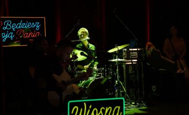 Na zdjęciu w widać mężczyznę grającego na perkusji, świeci na niego zielone światło. Znajdują się tam również dwaj gitarzyści oraz napisy led. 