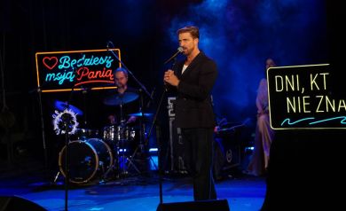 Na scenie muzyk śpiewający do mikrofonu. W tle  widać mężczyznę grającego na perkusji i kobietę z mikrofonem w ręku.Tło stanowią niebieskie światła oraz napis led. 