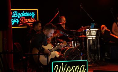 Na zdjęciu muzycy grający na instrumentach. W tle czerwone światła oraz napisy led. 