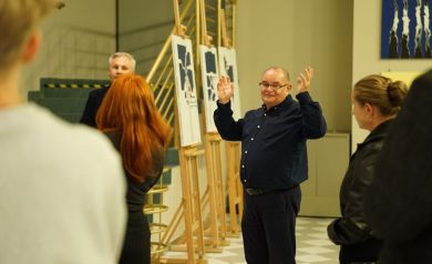 Na zdjęciu kilka osób. Jeden z mężczyzn w okularach gestykuluje rękoma. W tle widać obrazy stojące na sztalugach oraz wiszące na ścianie.