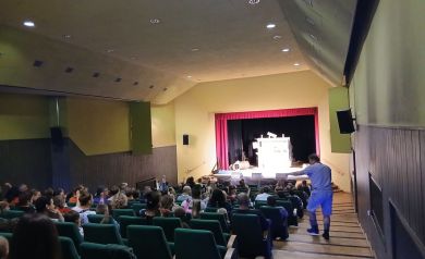 Na pierwszym planie publiczność i aktor idący po schodach, wskazujący ręką w stronę dzieci. 