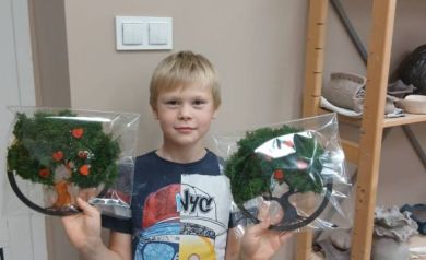 Na zdjęciu chłopczyk pokazujący do zdjęcia wykonane przez siebie dwa ,,Drzewka życia'. Obok niego regał z pracami ceramicznymi. 