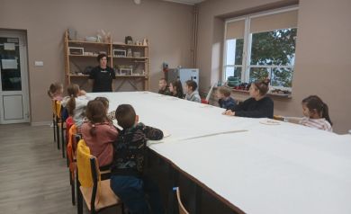 Grupa dzieci z prowadzącą warsztaty przy dużym stole nakrytym białym, papierowym obrusem. Widać również m.in. duże okno i regały z pracami ceramicznymi. 