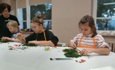 Na zdjęciu dzieci pracujące nad swoimi ,,Drzewkami życia' oraz kobieta prowadząca warsztaty pomagająca uczestnikom w wykonaniu swoich prac. 