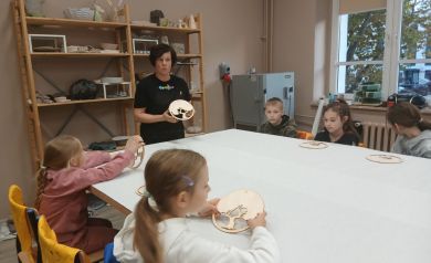 Na zdjęciu grupka dzieci biorący udział w warsztatach oraz prowadząca zajęcia. Dzieci siedzą przy stole nakrytym białym obrusem. W ich dłoniach lub na stole leży baza do wykonania ,,Drzewek życia'. Za prowadzącą znajdują się regały z pracami ceramicznymi z prawej strony widnieje okno, zaś w rogu sali jedna z maszyn wyposażająca pracownię ceramiczną. 