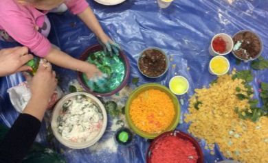 Na zdjęciu widać małe dziecko zanurzające ręce w misce z kolorową, zielono-niebieską masą podczas zabawy, otoczonej innymi miseczkami z barwionymi produktami spożywczymi. Obok znajduje się dorosła osoba pomagająca w zabawie oraz rozrzucone na folii materiały m. in. takie jak mąka, płatki i kolorowy makaron.