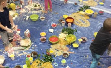 Na zdjęciu widać grupę małych dzieci bawiących się różnorodnymi materiałami spożywczymi rozłożonymi na dużej niebieskiej folii. To swobodna, kreatywna zabawa, podczas której dzieci mieszają, rozgniatają i eksplorują różne kolory oraz faktury.