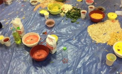 Na zdjęciu widać dzieci bawiące się na dużej niebieskiej folii, na której rozsypano i rozlano różnorodne produkty spożywcze jak mąka, płatki, makarony i kolorowe masy.