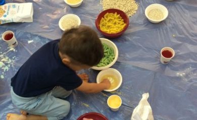 Na zdjęciu widać dziecko bawiące się na rozłożonej folii, dotykające misek z kolorowymi masami i produktami spożywczymi, takimi jak barwiony makaron czy płatki. Obok widać dorosłą osobę pomagającą w zabawie.
