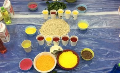 Na zdjęciu widać podłogę zabezpieczoną folią, na której rozmieszczono różnokolorowe produkty spożywcze w miseczkach i kubeczkach, takie jak płatki, kasze, barwione płyny czy zioła. 