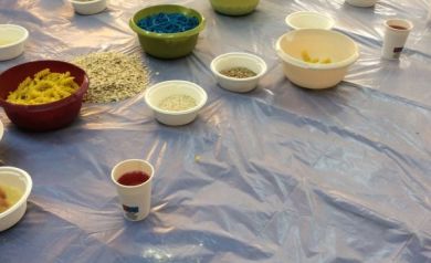 Na zdjęciu widać dziecko bawiące się na rozłożonej folii, dotykające misek z kolorowymi masami i produktami spożywczymi, takimi jak barwiony makaron czy płatki.