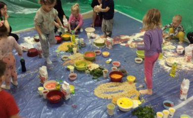 Na zdjęciu widać grupę dzieci bawiących się na dużej, zabezpieczonej folią powierzchni, gdzie rozsypano mąkę, płatki, makarony i inne kolorowe produkty spożywcze. Całość tworzy dynamiczną zabawę pod opieką dorosłych.