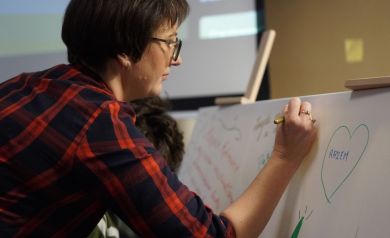 Kobieta w okularach i czerwonej sukience w kratkę pisze markerem na płótnie.