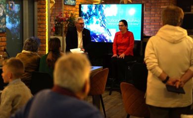 Na zdjęciu widać wernisaż wystawy, podczas którego dwoje prowadzących siedzi przed publicznością w przytulnym wnętrzu z ceglaną ścianą i ekranem w tle. Uczestnicy wydarzenia słuchają rozmowy, zajmując miejsca na krzesłach i stojąc w sali.