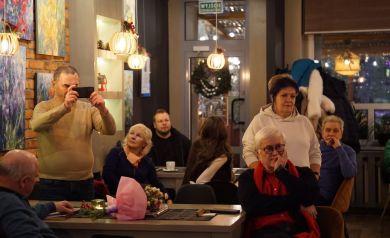 Na zdjęciu widać grupę ludzi zgromadzonych w kawiarni lub galerii, część siedzi przy stolikach, a jedna osoba robi zdjęcie telefonem. Atmosfera jest kameralna, a wnętrze ozdobione obrazami i ciepłym oświetleniem.