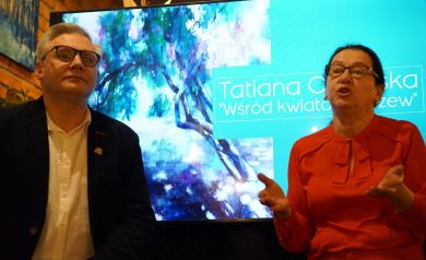 Na zdjęciu kobieta w czerwonej koszuli i okularach przemawia z rozłożonymi rękoma. Obok niej mężczyzna również w okularach, białej koszuli i ciemnej marynarce. W tle widać ekran z wyświetlaną grafiką. 