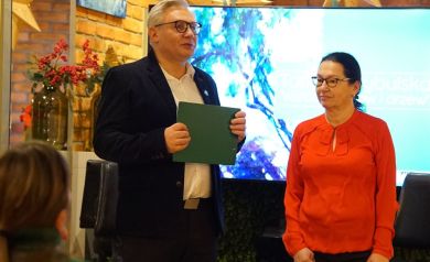 Na zdjęciu widać dwie osoby stojące obok siebie podczas wydarzenia w pomieszczeniu — mężczyzna trzyma zieloną teczkę i wygląda, jakby przemawiał, a kobieta stoi obok niego. W tle znajduje się ekran z grafiką oraz dekoracje na tle ceglanej ściany.