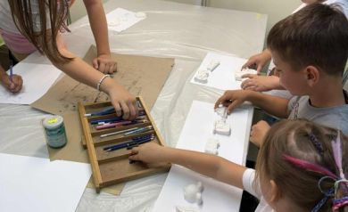 Przy stole pokrytym folią czwórka dzieci. Na białych kartkach leżą małe figurki. Na stole znajdują się również dwie skrzyni z kredkami, do jednej z nich sięgają dwie dziewczynki. 