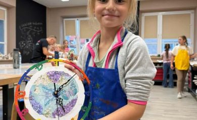 Dziewczyna w szaro-różowej bluzie prezentuje wykonany przez siebie zegar. 