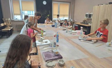 Dzieci uczestniczące w warsztatach ceramicznych. Znajdują się w pracowni i przy dużym stole wykonują prace ceramiczne. 