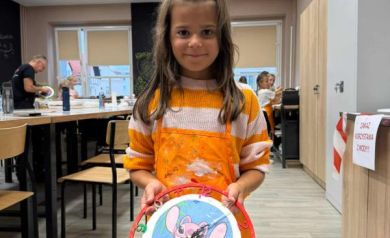 Dziewczynka ubrana w pomarańczowo-biały sweter i pomarańczowy fartuszek prezentuje swój zegar. W tle widać innych uczestników warsztatów i sale, gdzie obywały się zajęcia. 