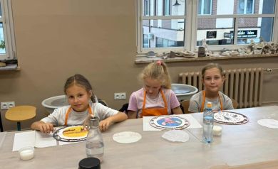 Trzy dziewczynki w pomarańczowych fartuszkach siedzące przy stole, przed nimi znajdują się wykonywane przez nie zegary. 