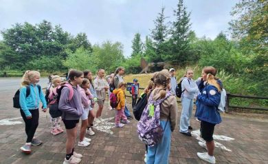 Grupka dzieci podczas wycieczki do JuraParku w Krasiejowie. W tle widać dużo zieleni i modele dinozaurów. 