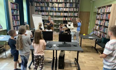 Na zdjęciu widoczna grupka dzieci i jedna kobieta. Kobieta mówi do dzieci, prowadzi zajęcia. Są w bibliotece (widać kilka regałów z książkami). Na zdjęciu są również czarne biurka, na których stoją komputery. 