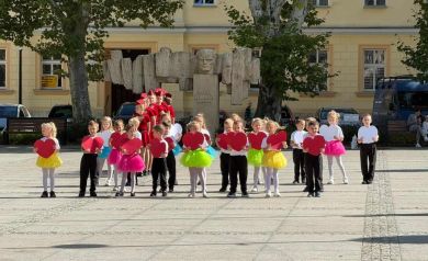 Na zdjęciu grupa przedszkolaków podczas swojego występu. Trzymają w dłoniach duże czerwone serca. Dziewczynki ubrane są w różnokolorowe spódniczki. Za nimi stoją w szeregu mażoretki, ubrane w czerwone sukienki.