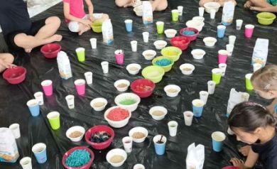 Podłoga jest wyłożona czarną folią. Na niej stoją różne kubki z kolorową zawartością. Wokół kubeczków w kółku siedzą dzieci biorące udział w zajęciach.