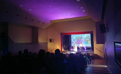 Na scenie występ kobiety i mężczyzny. Na pierwszym planie widać publikę. Światła oświetlają pomieszczenie na kolor fioletowy.