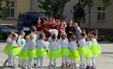 Na zdjęciu występ dzieci. Dziewczynki ubrane są w spódniczki jasnozielone. W tle widać mażoretki ubrane w czerwone stroje - stoją w szeregu.