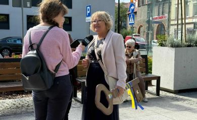 Kobieta udzielająca wywiadu, trzyma duży klucz w ręce. W tle widać budynki, znaki drogowe, donice z kwiatami oraz ławeczki.