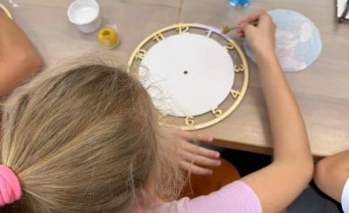 Dziewczynka ubrana w różową bluzkę, maluje na kolor fioletowy obręcz zegara. 