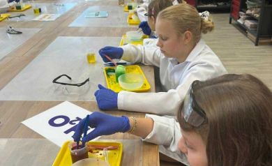 Dzieci w strojach chemików siedzą przy stole i  wykonują doświadczenia chemiczne. 