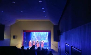 Na scenie zespół śpiewaczy podczas swojego występu. Na pierwszym planie widać publiczność. Światła oświetlają pomieszczenie na kolor niebieski.