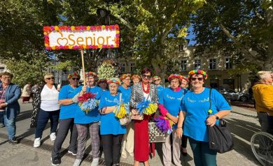 Grupa osób w większości ubranych w niebieskie koszulki z tabliczką z napisem ,,Senior+' pozuje do zdjęcia grupowego.