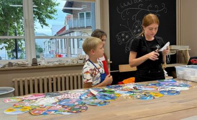 Na zdjęciu dwójka dzieci i prowadząca warsztaty. Na stole leżą różne wzory do udekorowania tarczy wykonywanych zegarów. 