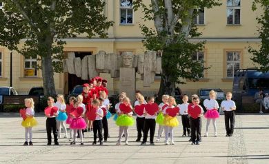 Na zdjęciu grupa przedszkolaków podczas swojego występu. Trzymają w dłoniach duże czerwone serca. Dziewczynki ubrane są w różnokolorowe spódniczki. Za nimi stoją w szeregu mażoretki, ubrane w czerwone sukienki.