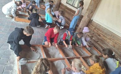 Dzieci przy trzech drewnianych skrzyniach, podzielonych na mniejsze skrzynki, przesiewają piasek.