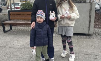Trójka uśmiechniętych dzieci trzyma znalezione torebki niespodzianki w kształcie zajączka. W tle hala sportowa na placu Stefana Żeromskiego.