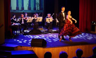 Para tancerzy przedstawia choreografię taneczną na scenie. W tle orkiestra grająca na instrumentach. 