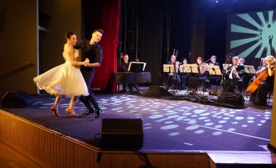 Para tancerzy przedstawia choreografię taneczną na scenie. Kobieta ma ubraną białą sukienkę, a mężczyzna czarną koszulę i kamizelkę. W tle orkiestra.
