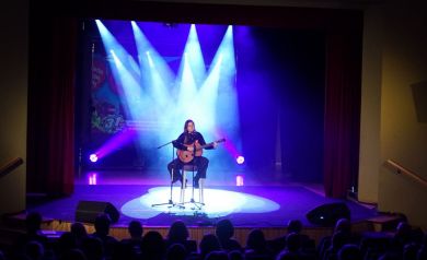 Na scenie kobieta grająca na gitarze w tle niebieskich świateł. 